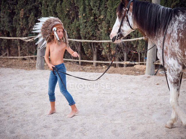 Menino calmo vestindo penas boné de guerra indiano e andando sem camisa na fazenda de areia, levando cavalo atrás — Fotografia de Stock