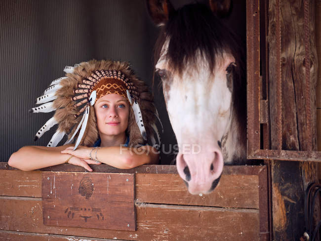 Mulher encantada em chapéu de pena indiana autêntica apoiada em cerca de madeira no estábulo com cavalo no rancho e olhando para longe — Fotografia de Stock