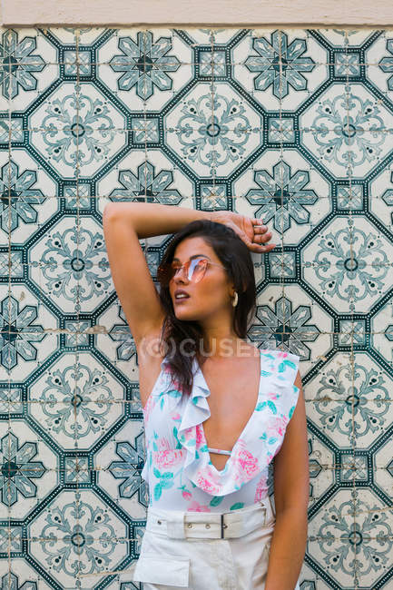 Peaceful gorgeous woman in trendy outfit and shiny sunglasses standing beside tiled exotic wall on scenic street — Stock Photo