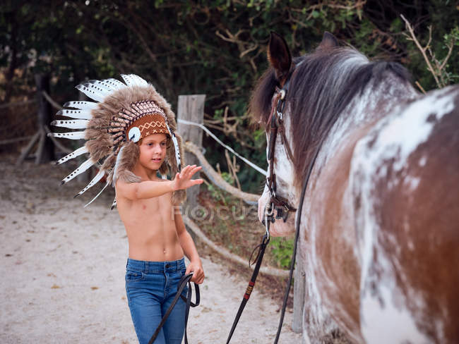 Calma ragazzo indossa cappello da guerra indiano piuma e camminare senza maglietta in fattoria sabbiosa, portando il cavallo dietro — Foto stock
