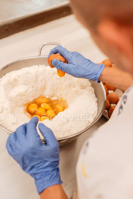 Сверху неузнаваемый кондитер в латексных перчатках разбивает свежие куриные яйца в миску с пшеничной мукой во время приготовления теста на кухне — стоковое фото