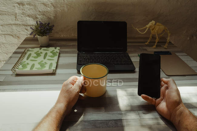 Homem irreconhecível com smartphone de navegação de copo de metal enquanto faz trabalho freelance na mesa com laptop e papelaria em casa — Fotografia de Stock