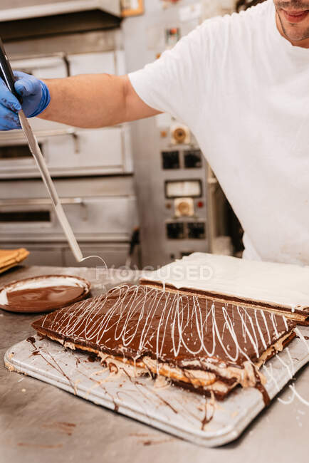 Homem de colheita em luva de látex e uniforme decorando delicioso bolo com redemoinhos de creme branco enquanto trabalhava na padaria — Fotografia de Stock