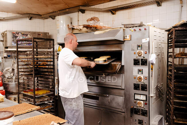 Man in uniform putting tray with raw cakes into hot oven while working in bakery — Stock Photo