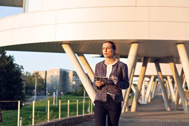 Serious businesswoman in elegant jacket browsing tablet and looking away while standing on city street on sunny day — Stock Photo