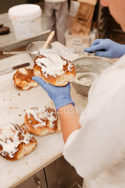 Зверху анонімний кондитер маскує м'який солодкий крем на свіжій булочці під час роботи в хлібобулочних виробах — стокове фото