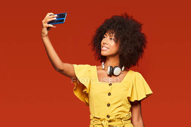 Mujer afroamericana que lleva auriculares en el cuello y que se toma selfie con teléfono móvil sobre fondo rojo vivo. - foto de stock