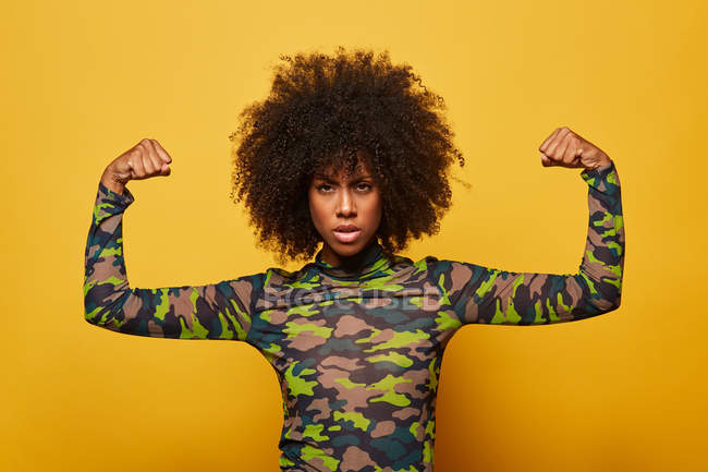 Une Afro-Américaine en chemise de camouflage portant des biceps sur fond jaune regarde la caméra — Photo de stock