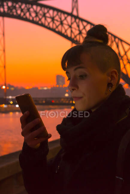 Trendy modern female with hair bun and piercing browsing smartphone while standing on embankment on blurred background of river and sunset sky in city — Stock Photo