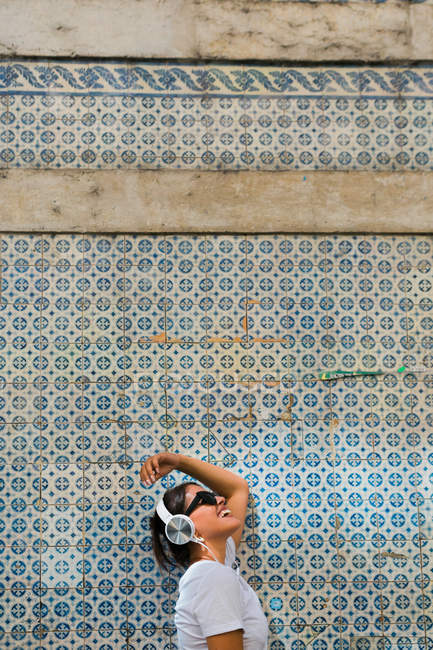 Беззаботная красивая женщина в повседневной одежде и наушниках, стоящая на синей мозаичной стене здания на городской улице — стоковое фото