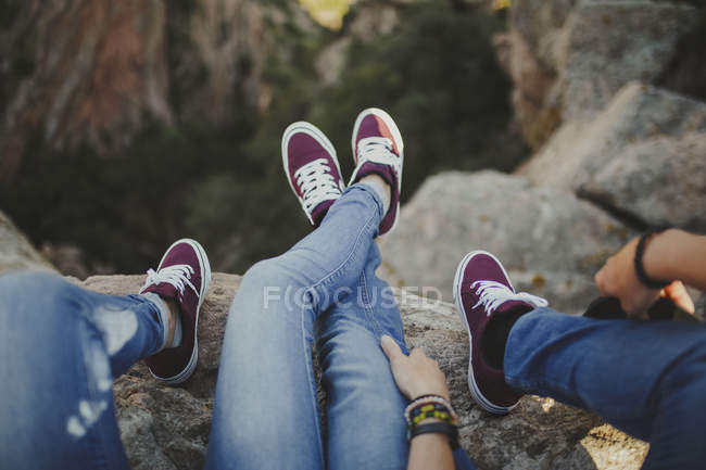 Cruza desde arriba los pies relajados pareja en un equipo de denim y zapatillas tumbadas en abrazo en el acantilado disfrutando de las vistas. - foto de stock