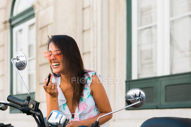 Carefree beautiful woman in sunglasses putting on bright lipstick while looking at motorbike mirror on street — Stock Photo