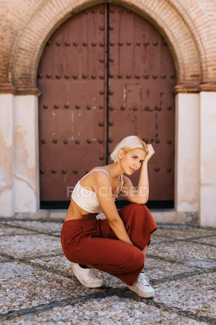 Atractiva mujer joven en traje elegante agacharse, mirando hacia otro lado contra el edificio antiguo con puerta de mala muerte en la calle del casco antiguo - foto de stock