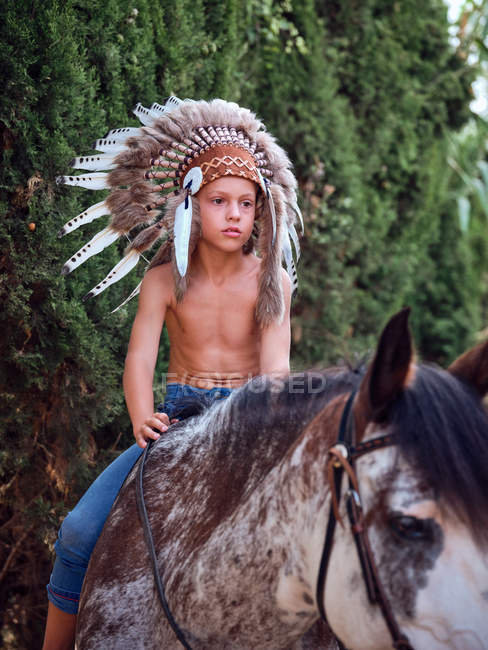 Serious boy in authentic Indian feather hat riding horse in park — Stock Photo