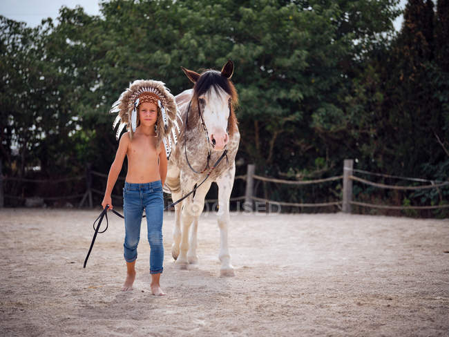 Calm kid wearing feather Indian war bonnet and walking shirtless on sandy farm, leading horse behind — Stock Photo