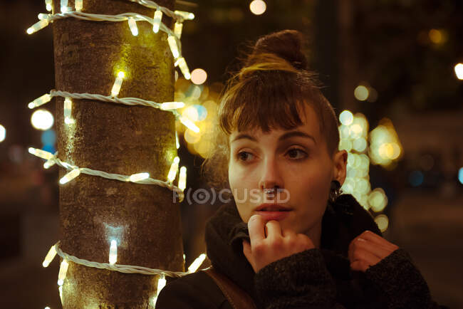 Pretty young female with hair bun and piercing looking away while standing near tree trunk decorated with fairy lights on city street in evening — Stock Photo