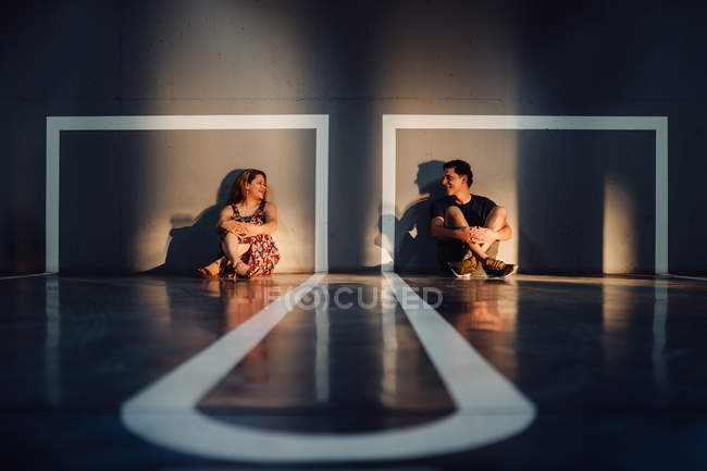 Charming couple sitting on floor of marked playground at sunlight and looking with tender tender. - foto de stock