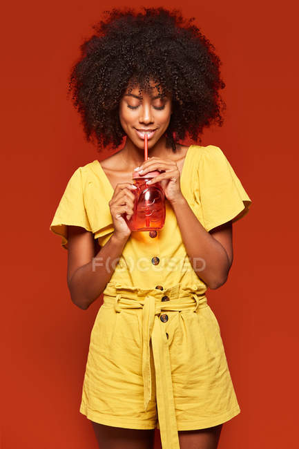 Femme afro-américaine rêveuse aux cheveux frisés tenant un pot rouge avec de la paille et savourant une boisson sur fond rouge — Photo de stock