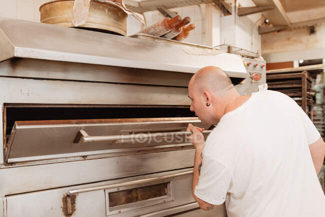Adult overweight man with bald head peeking inside professional oven while working in bakery — Stock Photo