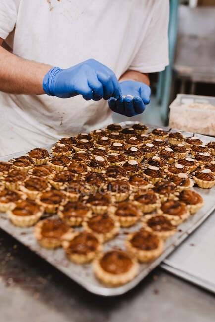 Неузнаваемый человек в латексных перчатках кладет орехи поверх сладких пирожных во время работы в пекарне — стоковое фото