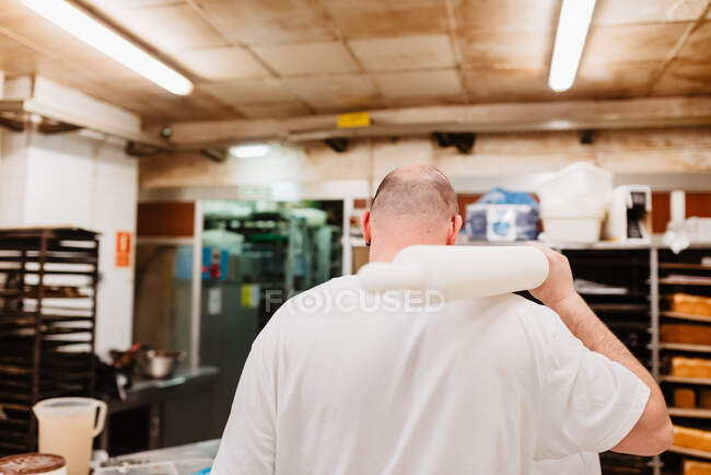 Back view of bald overweight man in white uniform holding rolling pin on shoulder while working in bakery — Stock Photo