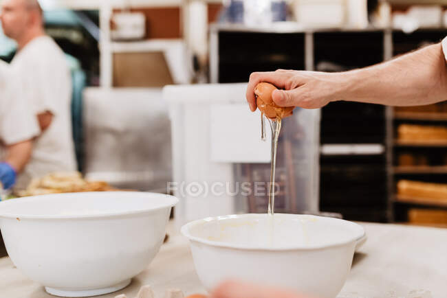 D'en haut confiseur méconnaissable dans des gants en latex briser les œufs de poulet frais dans un bol avec de la farine de blé tout en préparant la pâtisserie dans la cuisine — Photo de stock