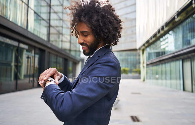 Веселый африканский офисный работник с вьющимися волосами, стоящий на улице и смотрящий на часы на размытом фоне — стоковое фото