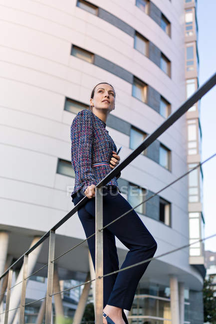 Low angle of serious female manager in elegant outfit leaning on railing and looking away — Stock Photo
