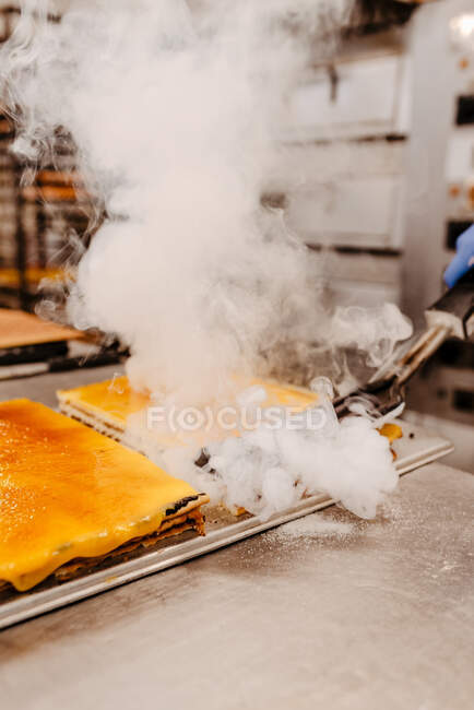Unrecognizable cook melting cake topping with hot tool emitting smoke over table in bakery — Stock Photo