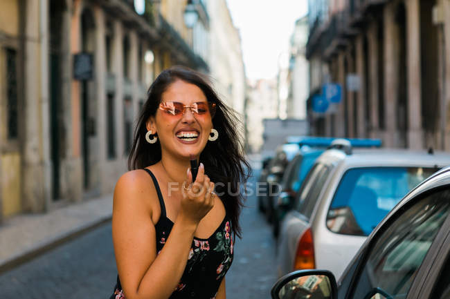 Femme en lunettes de soleil et robe portant du rouge à lèvres tout en regardant la caméra, debout près du miroir de voiture sur la rue ensoleillée — Photo de stock