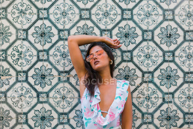 Peaceful gorgeous woman in trendy outfit and shiny sunglasses leaning on tiled exotic wall with closed eyes on scenic street — Stock Photo