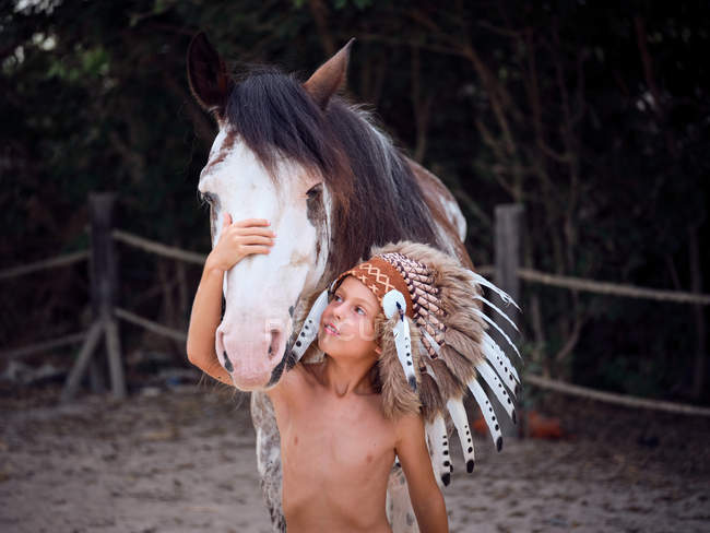 Tranquil child wearing traditional Indian war bonnet, bonding with horse stallion on blurred background — Stock Photo