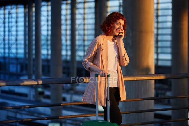 Счастливая рыжая молодая женщина с чемоданом разговаривает по мобильному телефону на станции вечером — стоковое фото