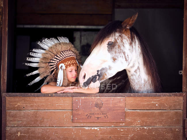 Delighted kid in authentic Indian feather hat leaning on wooden fence in stable with horse on ranch and looking away — Stock Photo