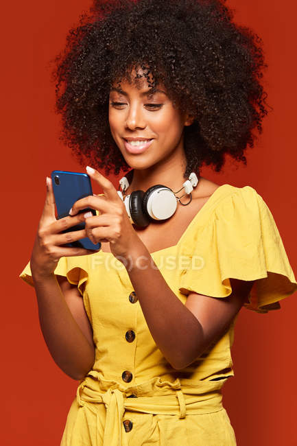 Mulher americana africana feliz moderna vestindo fones de ouvido no pescoço e usando telefone celular no fundo vermelho vívido — Fotografia de Stock