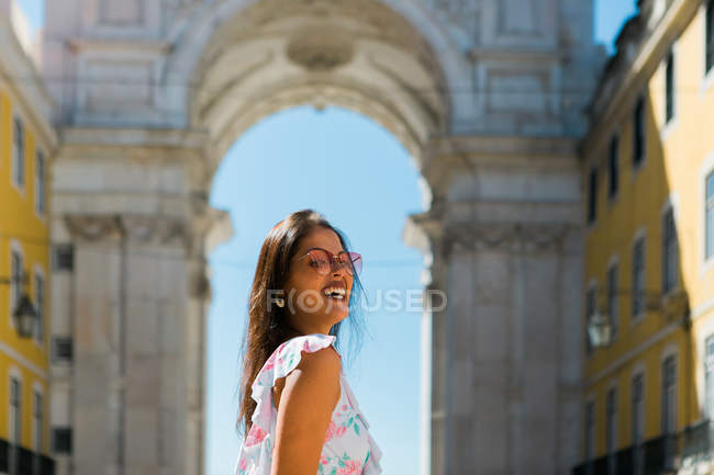 Joven mujer alegre en gafas de sol de pie junto al majestuoso arco en la calle de la ciudad en Lisboa, Portugal - foto de stock