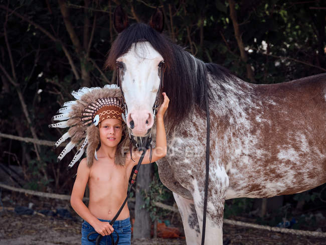 Delighted kid in Indian feather war bonnet caressing horse on ranch and looking at camera — Stock Photo