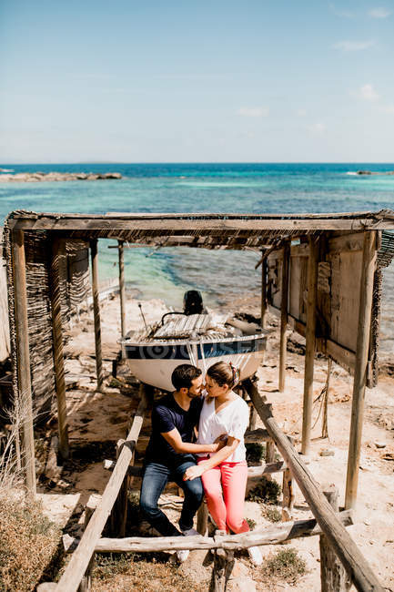 Aimant couple câlins près du bateau amarré à terre sous la verrière en bois sur la plage en été — Photo de stock