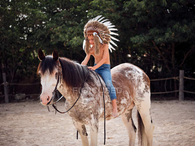 Garçon sérieux en authentique chapeau de plume indien équitation cheval dans le parc — Photo de stock