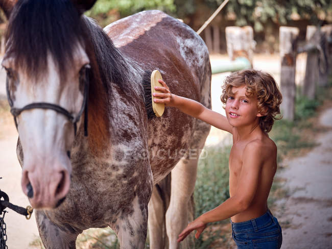 Conteúdo menino em jeans grooming cavalo com escova no rancho e olhando para a câmera no fundo borrado — Fotografia de Stock
