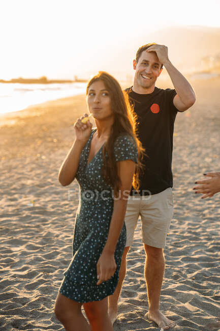 Ein schönes Mädchen, das Pommes frites isst, und ihr Freund, der am Strand in die Kamera lächelt und die Sonne hinter sich hat — Stockfoto
