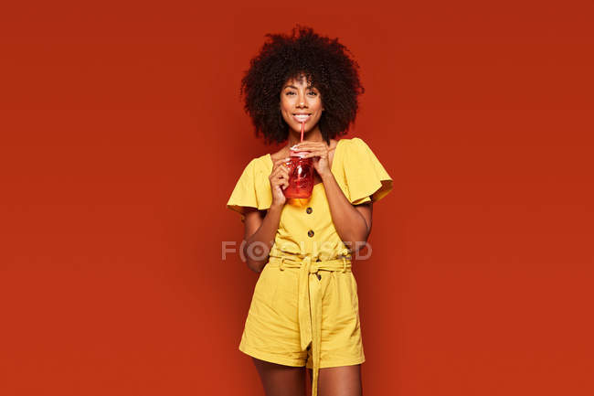 Cheerful African American woman holding red jar with straw and enjoying beverage on red background looking at camera — Stock Photo