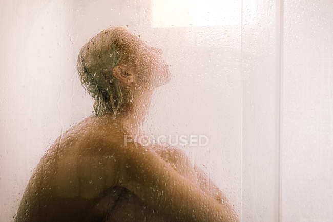 Vista lateral da jovem mulher nua tomando banho atrás de divisória transparente molhada no banheiro — Fotografia de Stock