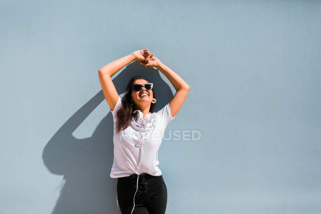 Carefree beautiful woman in casual outfit and headphones on neck standing with hands up beside blue wall of building on city street — Stock Photo