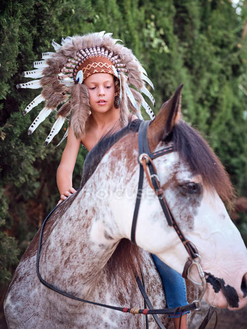 Garçon sérieux en authentique chapeau de plume indien équitation cheval dans le parc — Photo de stock