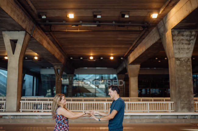 Seitenansicht des schönen Paares, das Zeit miteinander verbringt und Händchen klammerndes Spiel unter großer Brücke im Abendlicht auf der Stadtstraße spielt — Stockfoto