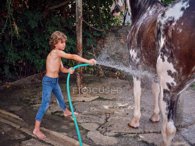 Seitenansicht des barfüßigen Jungen, der Hengst mit Süßwasser auf Bauernhofterrasse abspritzt — Stockfoto