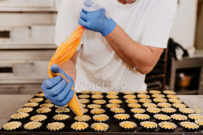 Анонимный кондитер в латексных перчатках сжимает вкусное сладкое желе из сумки в маленькие кондитерские коробочки на размытом фоне пекарской кухни — стоковое фото