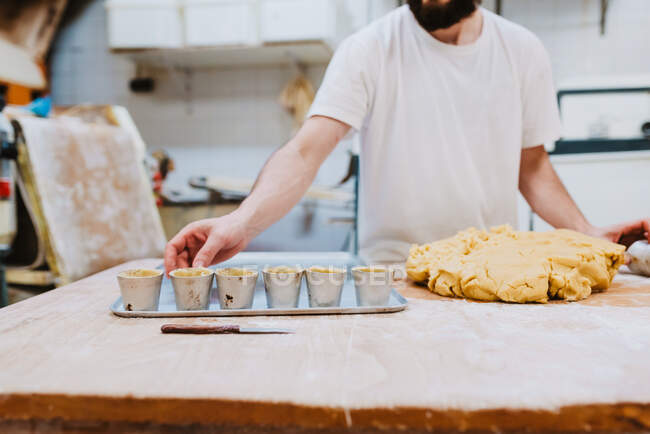 Homem barbudo da colheita na t-shirt branca que põe a massa fresca em copos ao fazer a pastelaria na cozinha da padaria — Fotografia de Stock