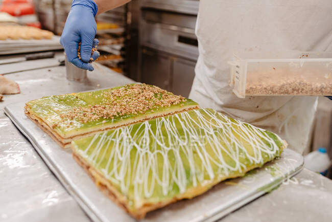 Unbekannter Konditor im Handschuh dekoriert leckeren frischen Kuchen mit Gebäckkrümeln während er in der Küche der Bäckerei arbeitet — Stockfoto